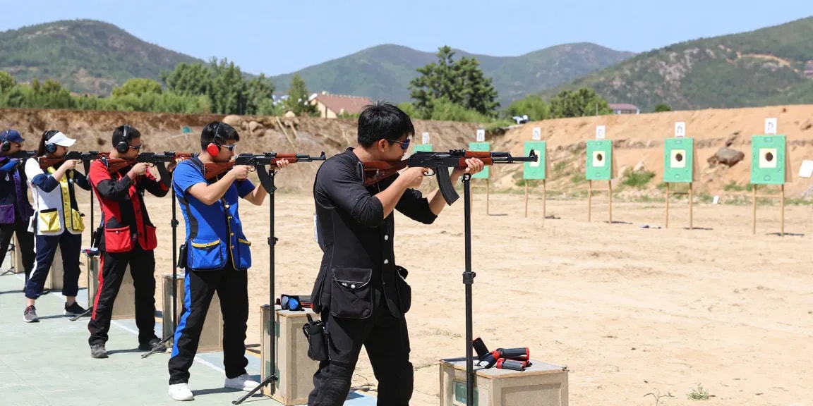 Group of enthusiasts practicing with an 8k47-gun at an outdoor shooting range, showcasing safety gear and mountainous backdrop.