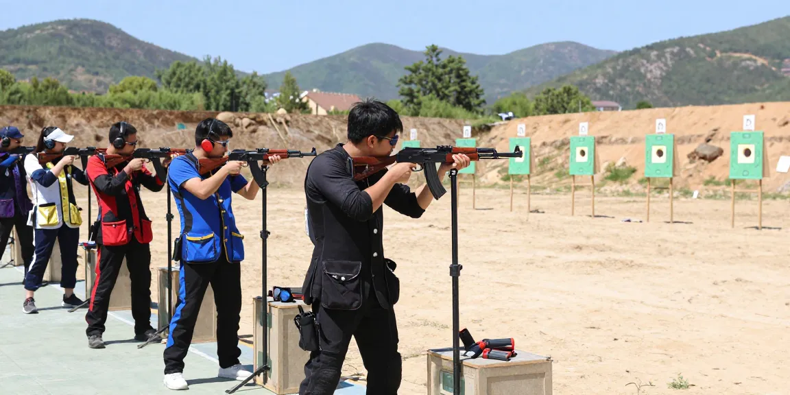 Group of enthusiasts practicing with an 8k47-gun at an outdoor shooting range, showcasing safety gear and mountainous backdrop.